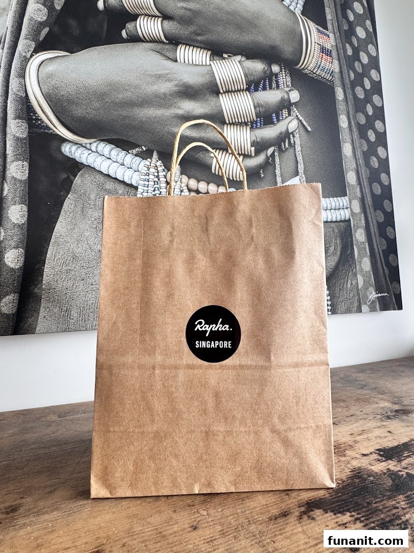A brown paper shopping bag with a black 'Rapha. Singapore' logo sits on a wooden surface.