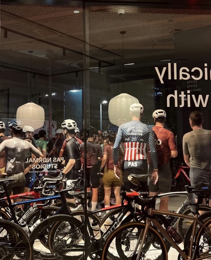 Cyclists and high-end bicycles are displayed inside Pas Normal Studios, viewed through a glass window.