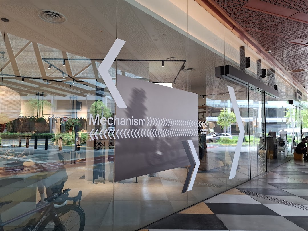The glass facade of Pas Normal Studios Singapore, featuring 'Mechanism' branding and a view of the interior.