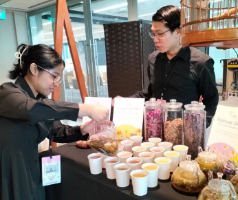 Two staff members serve tea samples from a display with various tea blends and brewing pots.