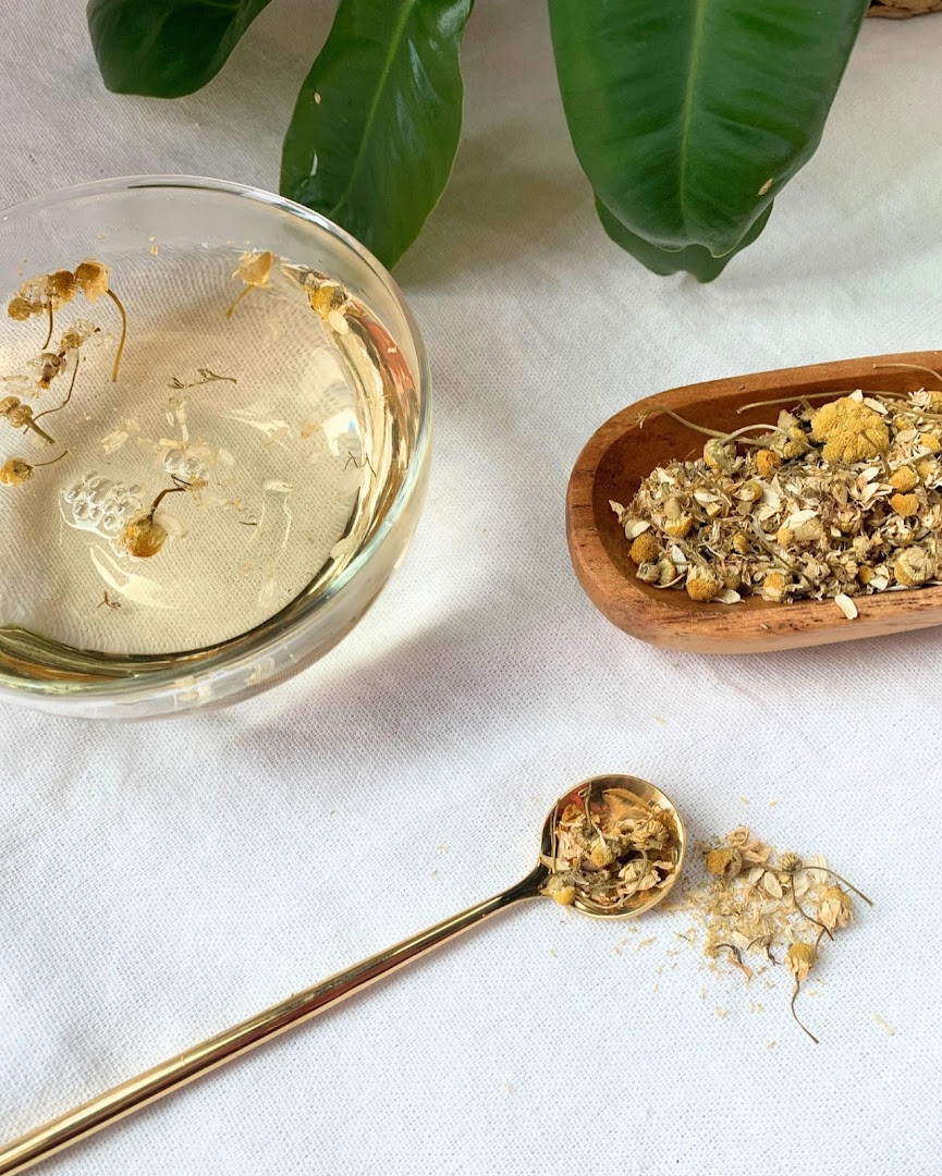 A clear glass bowl of chamomile tea, dried chamomile flowers, and a golden spoon on a white surface.