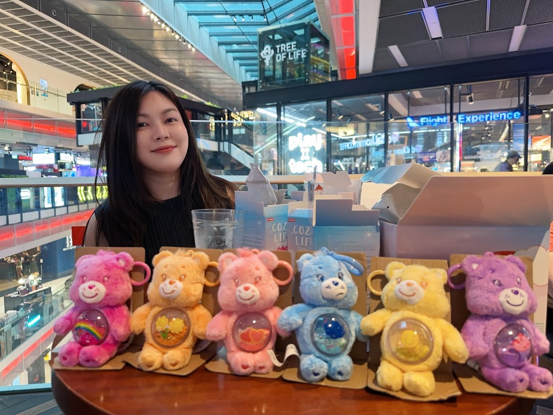 A person smiles behind a display of colorful Care Bear plush keychains inside a vibrant shopping mall.