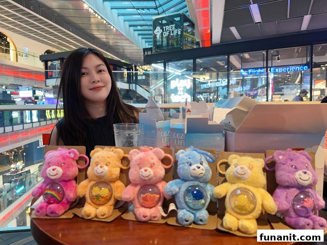 A person smiles behind a display of colorful Care Bear plush keychains inside a vibrant shopping mall.