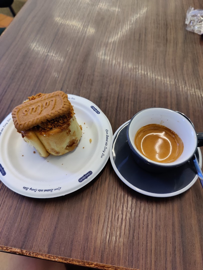 A close-up of a coffee cup and a Cinnabon pastry topped with a Lotus Biscoff cookie on a wooden table.