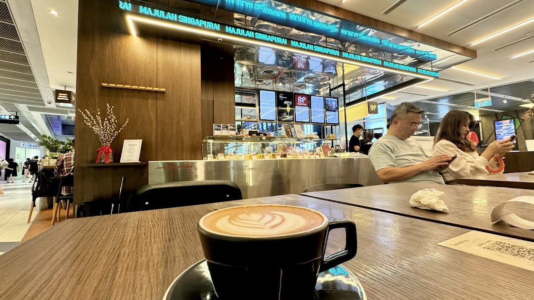 A warm latte with latte art sits on a wooden table inside PPP Coffee, with customers and the counter in the background.