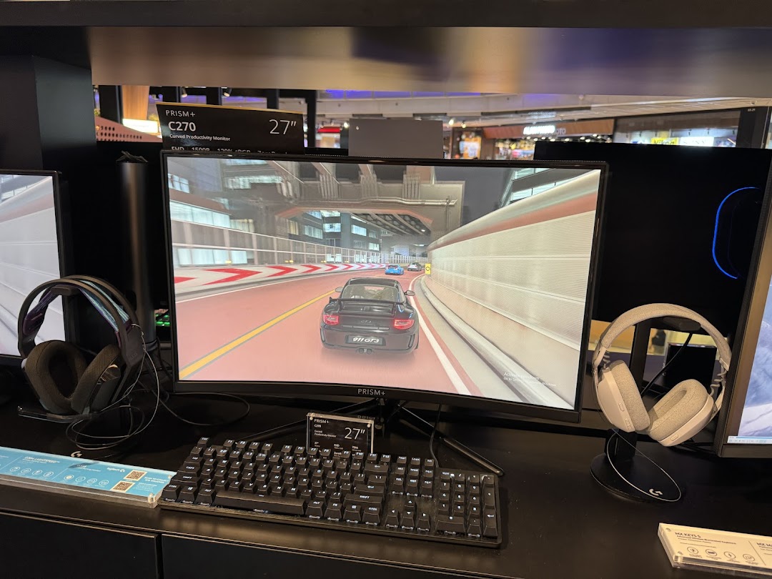 A curved monitor displaying a racing game, flanked by gaming headphones and a keyboard, showcasing tech products.