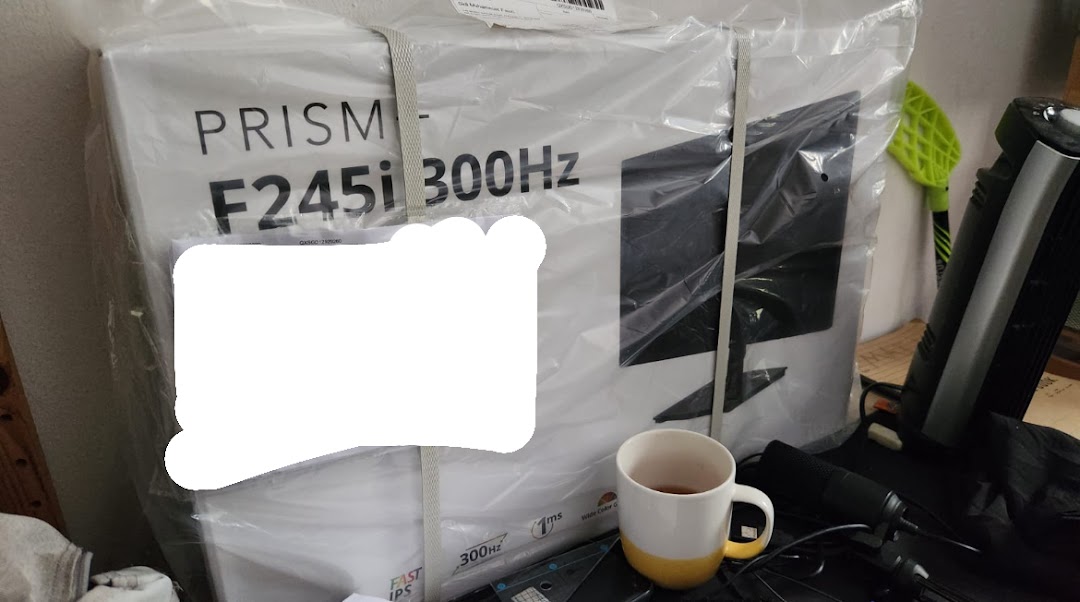 A large monitor box from PRISM Funan, still in its plastic packaging, with a mug nearby.