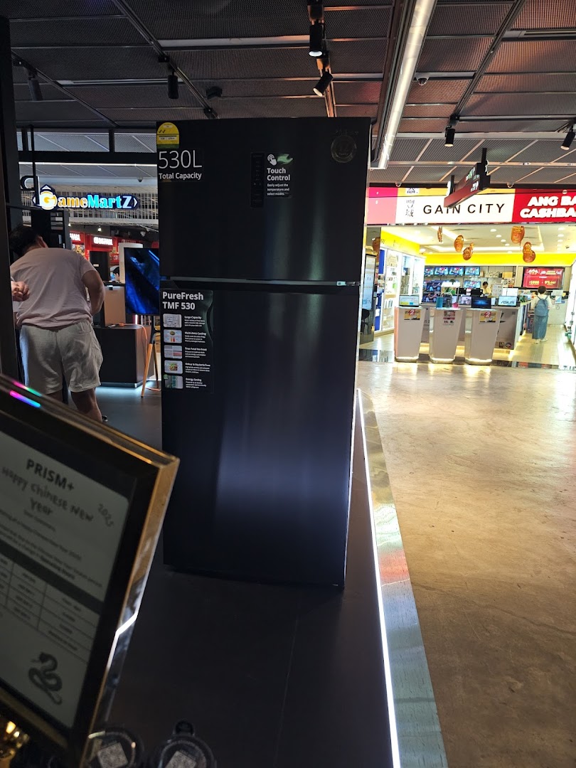 A large black refrigerator on display inside PRISM Funan, an electronics store, with other shops visible in the background.