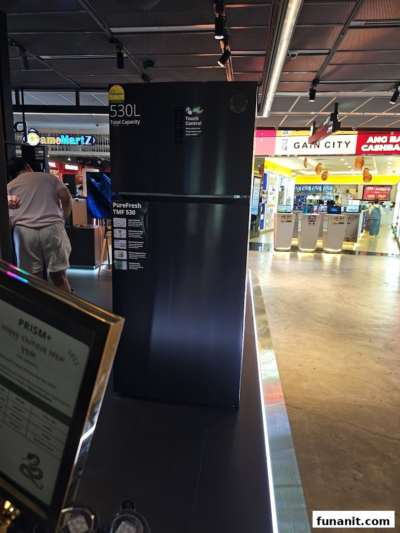 A large black refrigerator on display inside PRISM Funan, an electronics store, with other shops visible in the background.