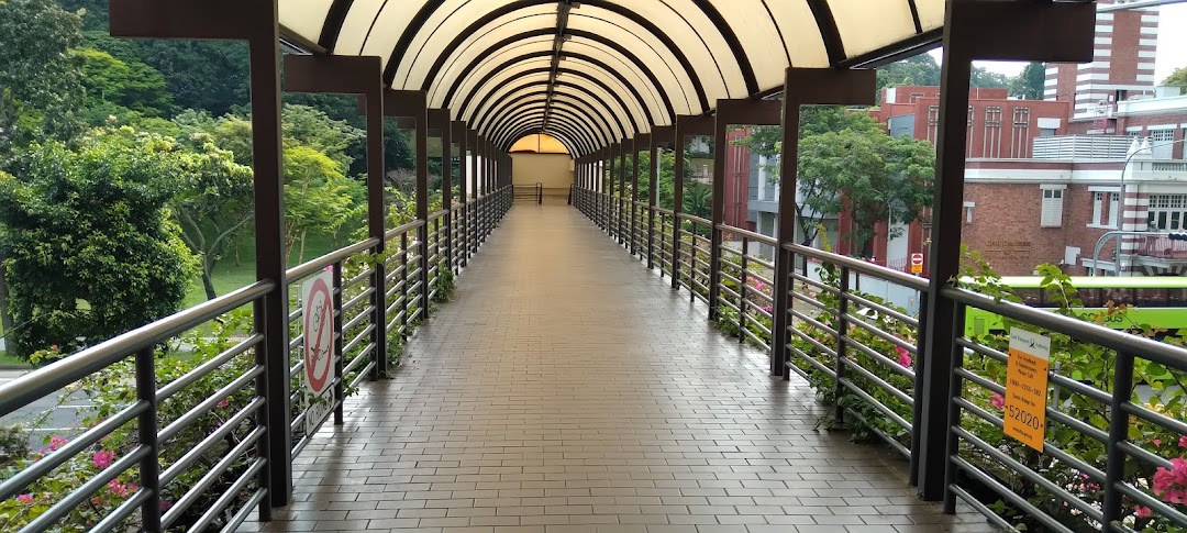 A long, covered pedestrian walkway with a vaulted roof, surrounded by lush greenery and distant buildings.
