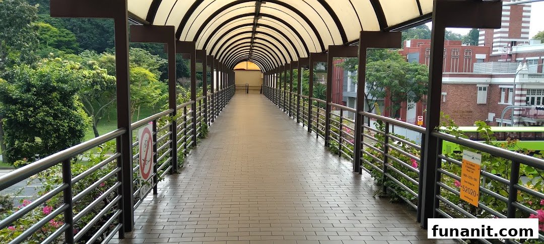 A long, covered pedestrian walkway with a vaulted roof, surrounded by lush greenery and distant buildings.