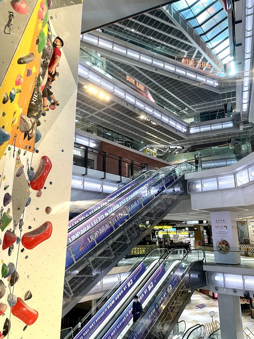 A person rock climbing on an indoor wall next to escalators in a modern multi-level shopping mall.