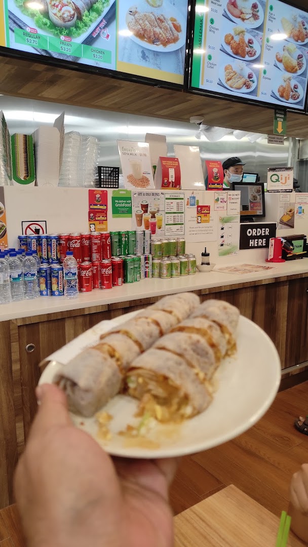 A person holds spring rolls in a food stall with digital menu boards displaying various dishes.