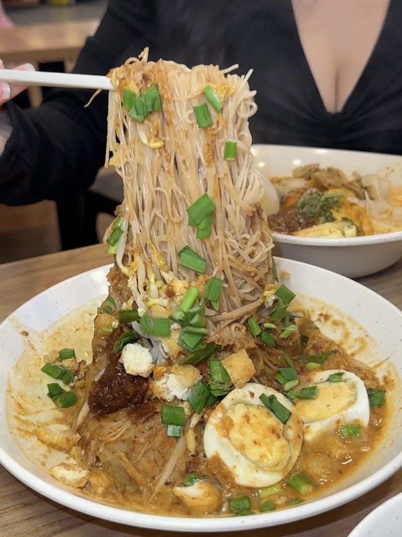 A close-up of a person lifting flavorful noodles from a rich, saucy dish with eggs and sprouts.