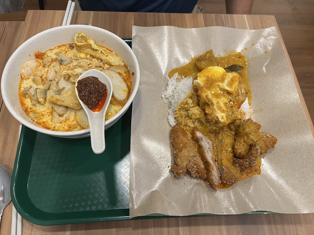 A vibrant bowl of laksa and a plate of curry chicken rice with egg, served on a wooden table.