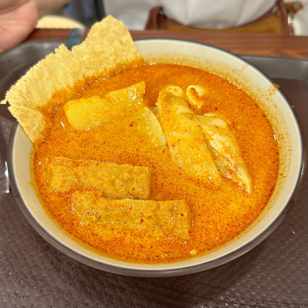 A vibrant bowl of curry or laksa with tofu, fish cakes, and a crispy cracker, served on a tray.