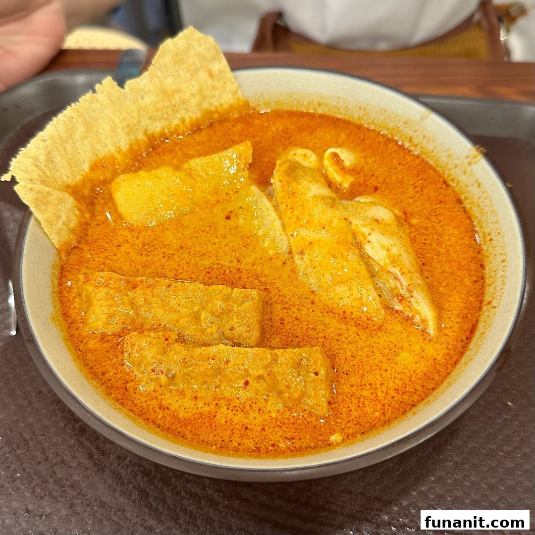 A vibrant bowl of curry or laksa with tofu, fish cakes, and a crispy cracker, served on a tray.