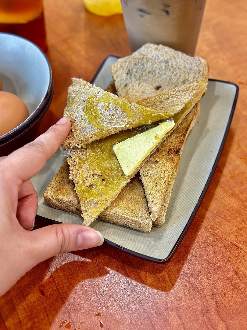A hand reaches for a plate of traditional kaya toast with butter, served with eggs and drinks.