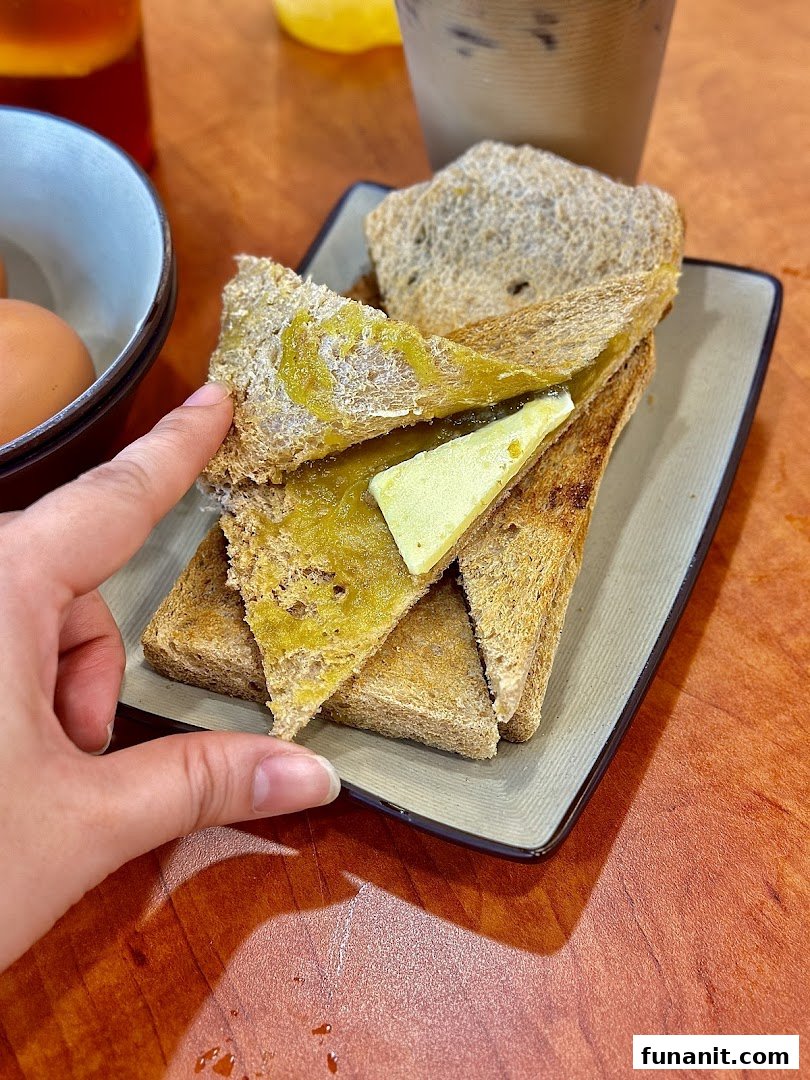 A hand reaches for a plate of traditional kaya toast with butter, served with eggs and drinks.