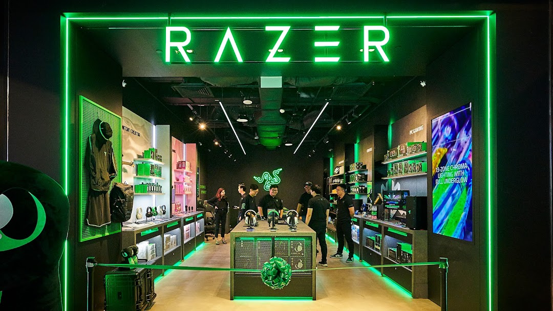 The RazerStore Funan interior features vibrant green lighting, displaying gaming peripherals and electronics with staff present.