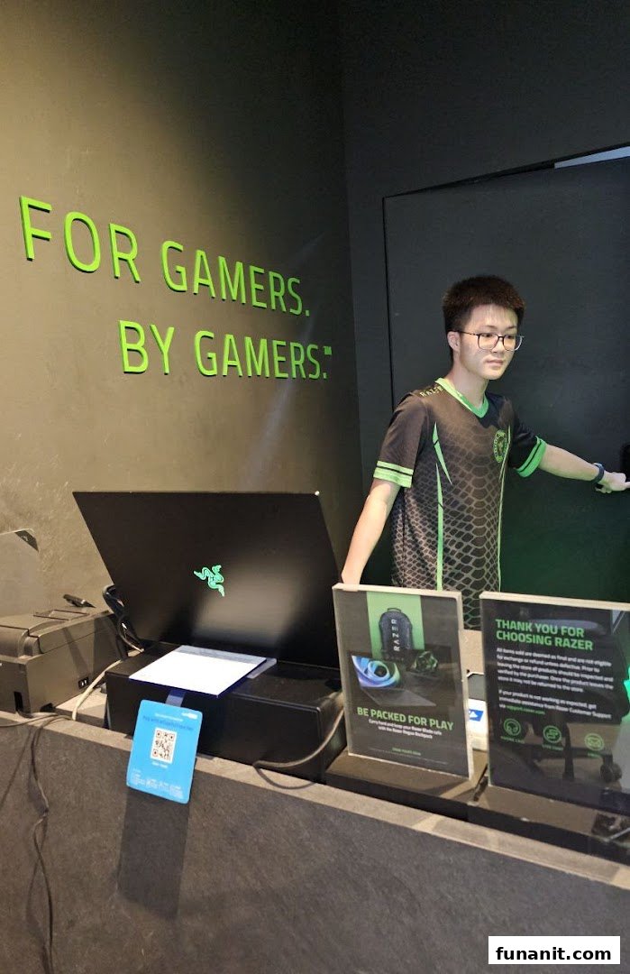 A Razer store employee stands behind a counter with a laptop and promotional signs.
