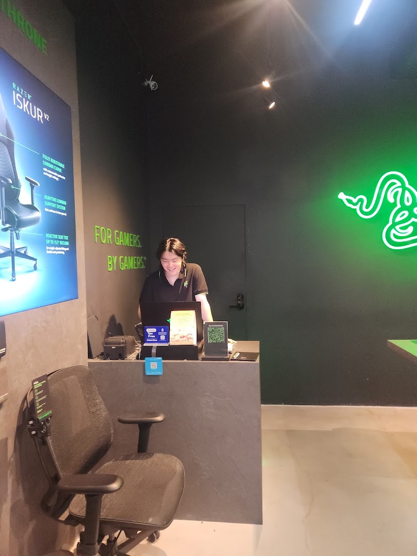 An interior view of RazerStore Funan, featuring a staff member, a gaming chair display, and the brand's iconic green neon logo.