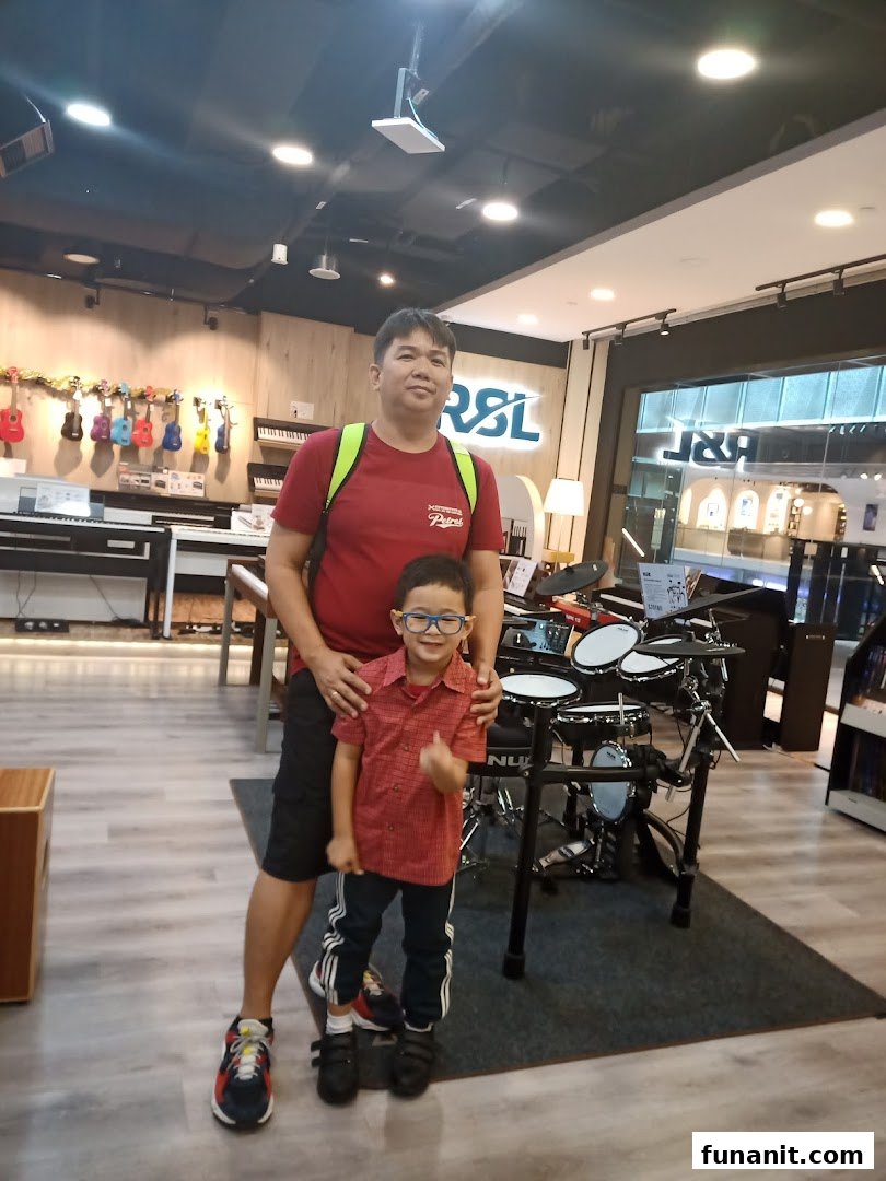 A man and a child pose in a music instrument shop featuring drums, keyboards, and ukuleles.