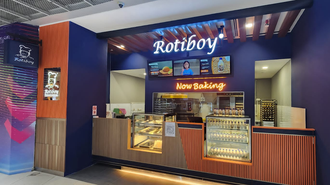A vibrant Rotiboy shop front with a counter, display cases, and 'Now Baking' sign.