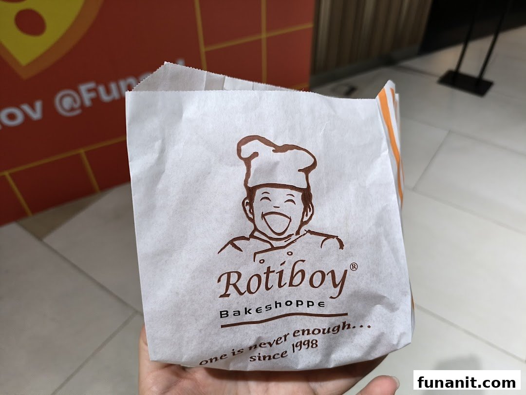 A hand holds a Rotiboy Bakeshoppe paper bag featuring its iconic logo and slogan.