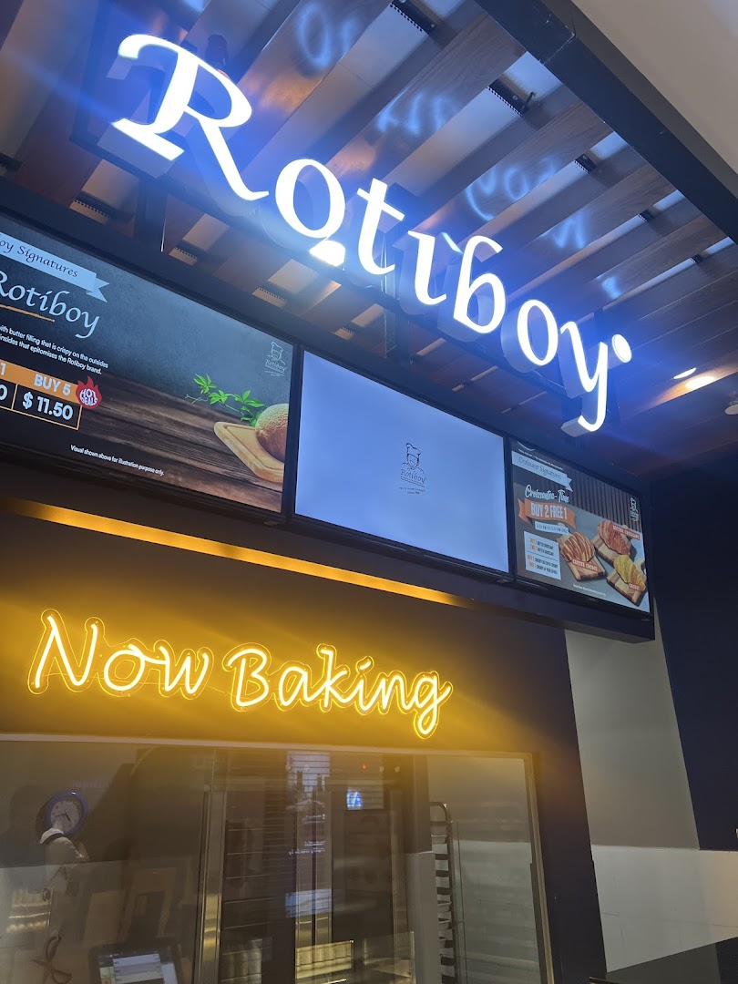 The illuminated Rotiboy shop front features its iconic sign, digital displays, and a 'Now Baking' neon sign.