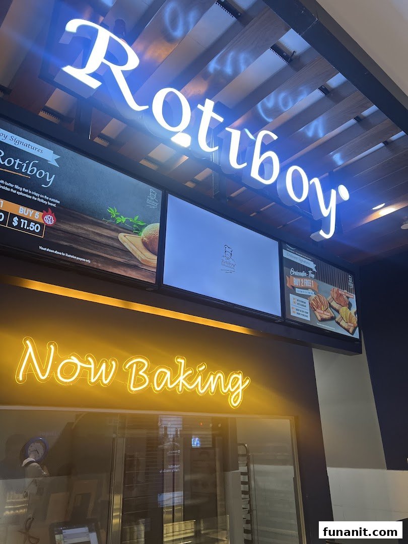 The illuminated Rotiboy shop front features its iconic sign, digital displays, and a 'Now Baking' neon sign.
