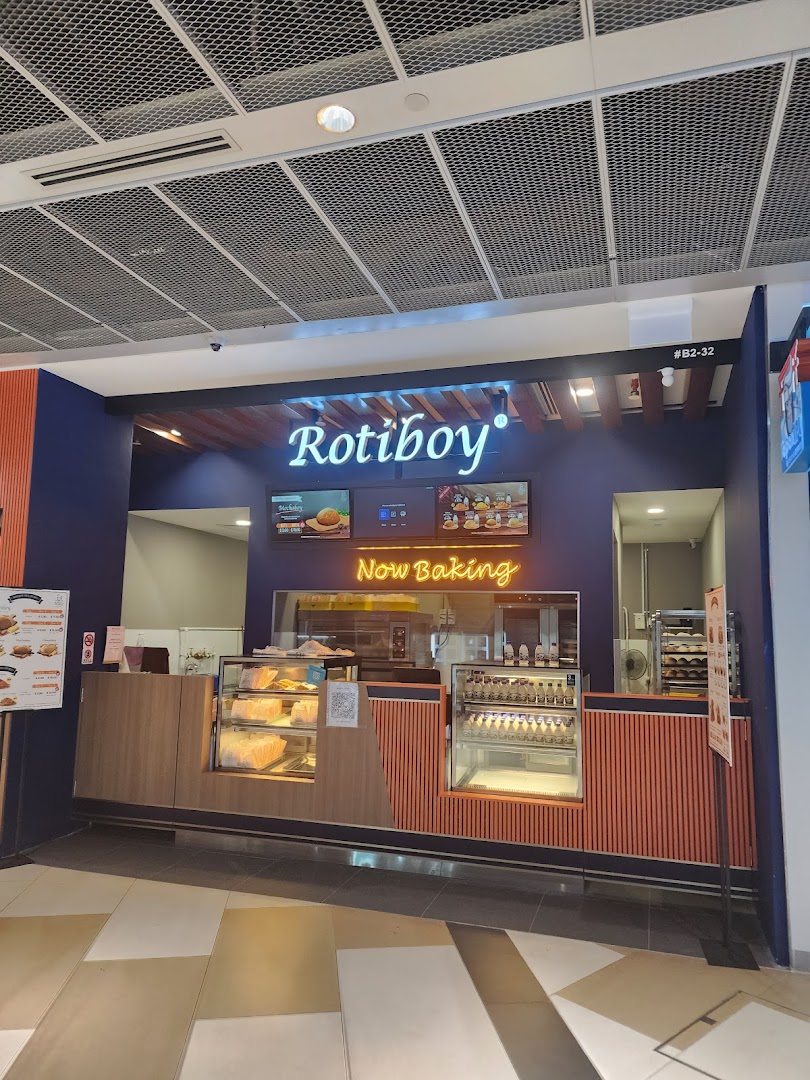 The storefront of Rotiboy bakery, featuring its illuminated sign, display counters, and a 'Now Baking' neon sign.