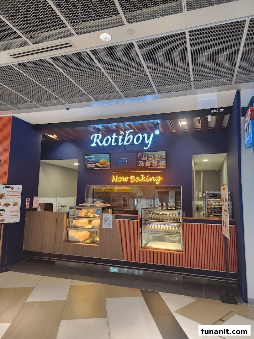 The storefront of Rotiboy bakery, featuring its illuminated sign, display counters, and a 'Now Baking' neon sign.