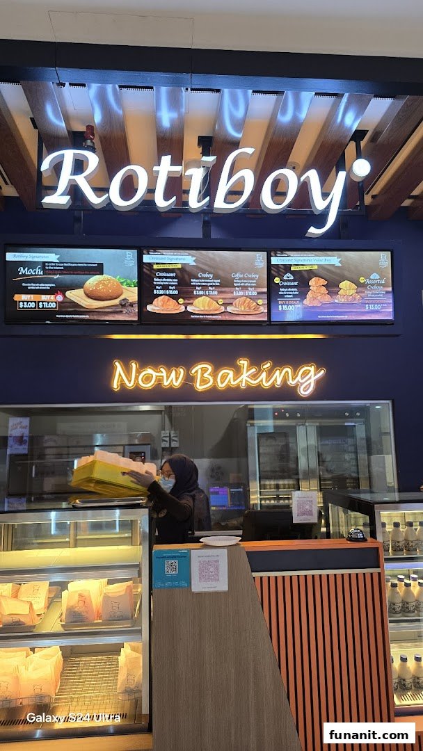 A Rotiboy shop interior featuring digital menu boards, a staff member, and baked goods display.
