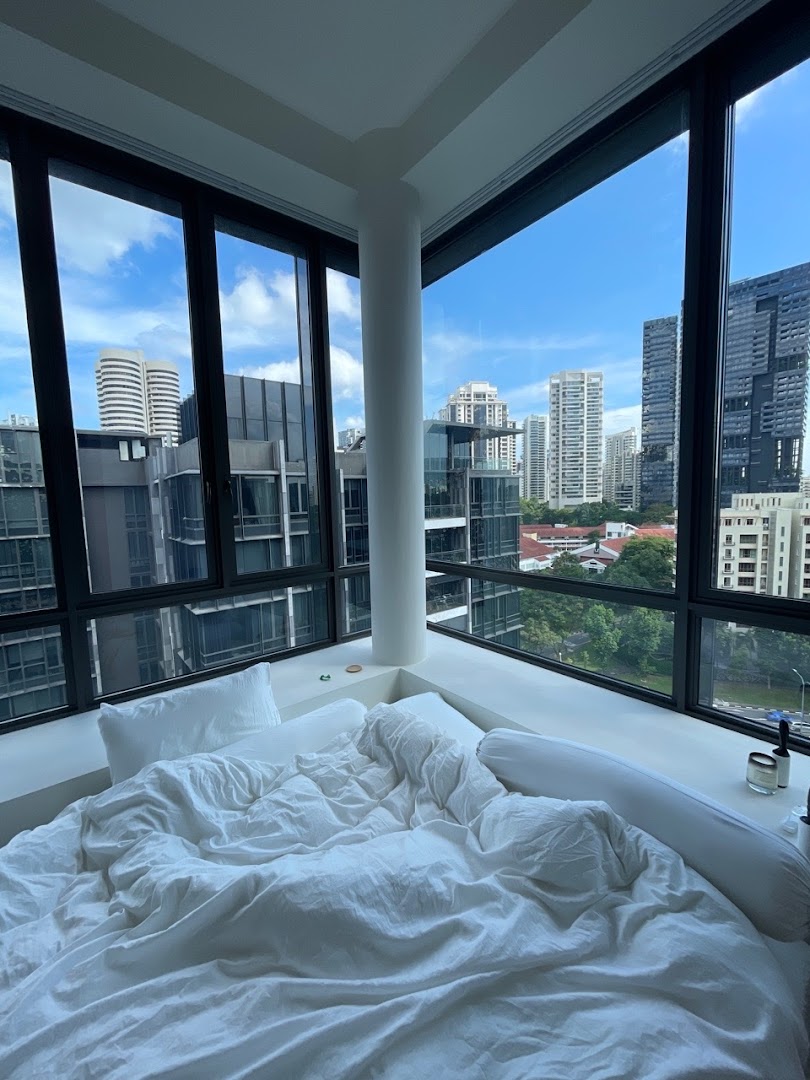 A cozy bed with white linens positioned by large windows overlooking a modern city skyline.