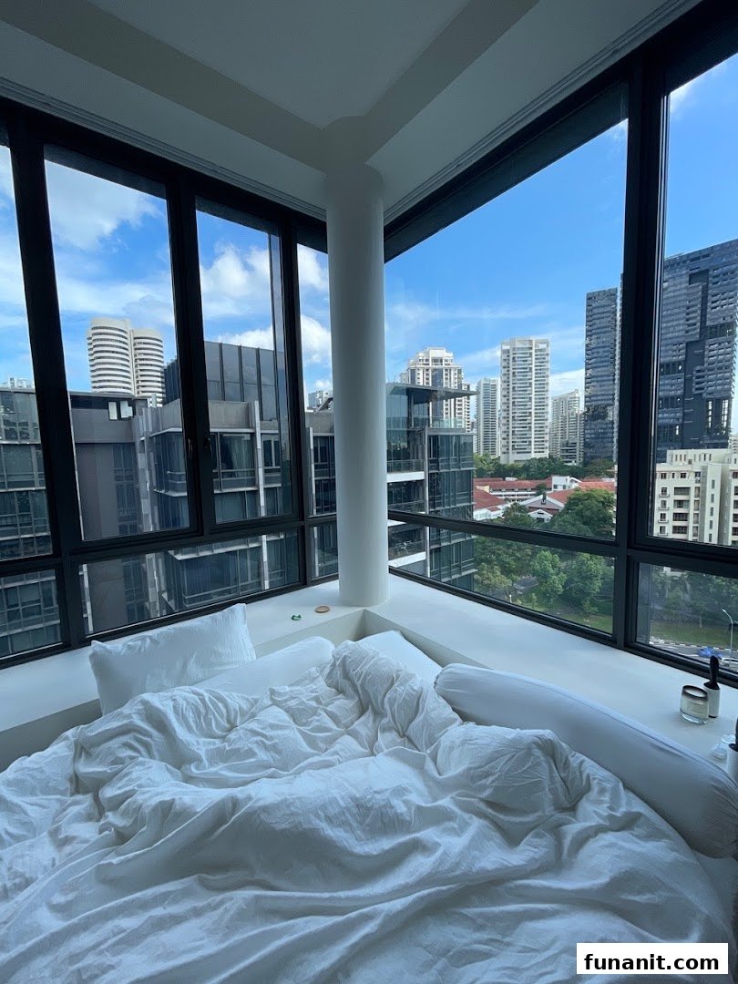 A cozy bed with white linens positioned by large windows overlooking a modern city skyline.