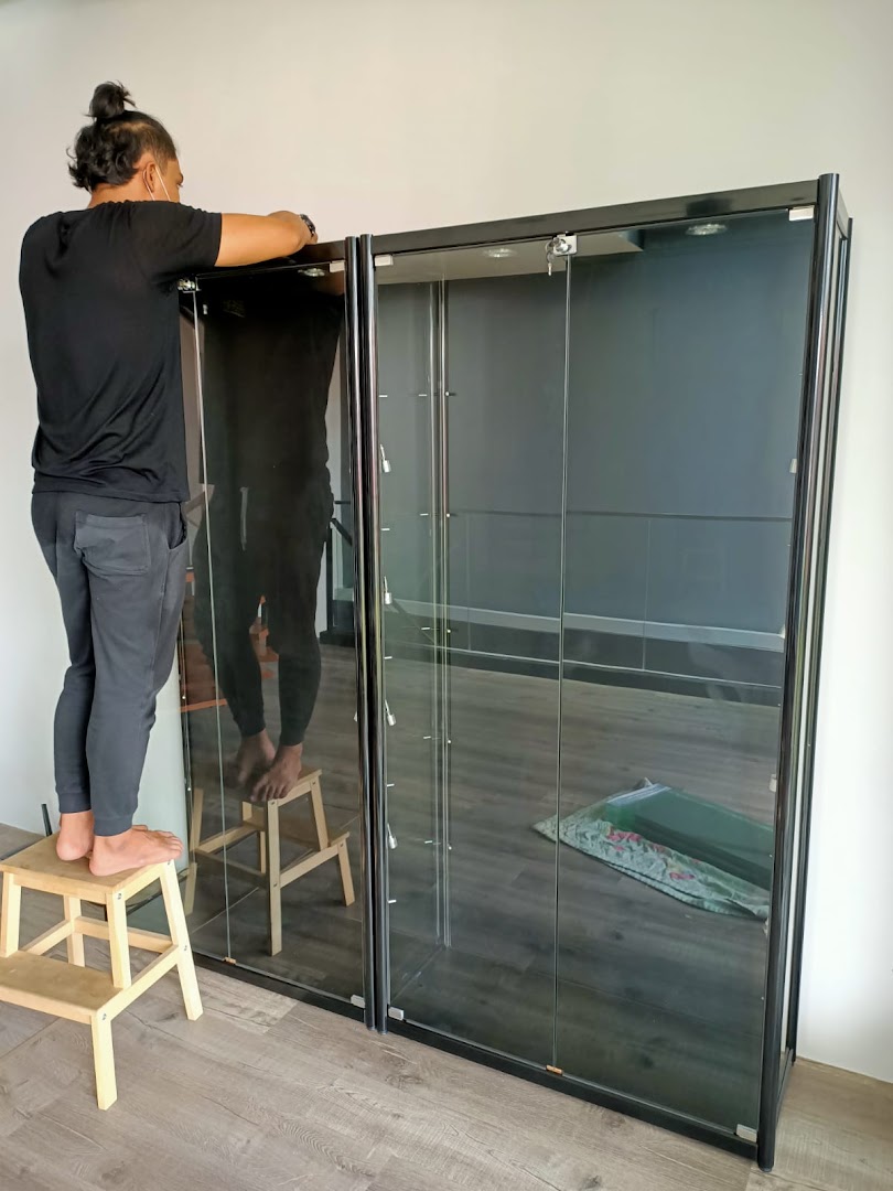 A person assembling or adjusting a black-framed glass display cabinet for toys or collectibles.