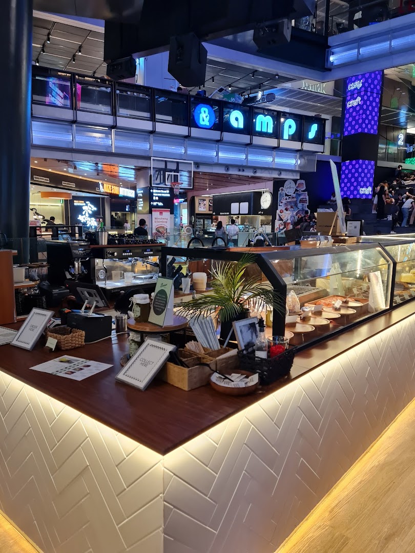 A vibrant coffee shop counter with a display case, plants, and various items, set within a bustling mall.