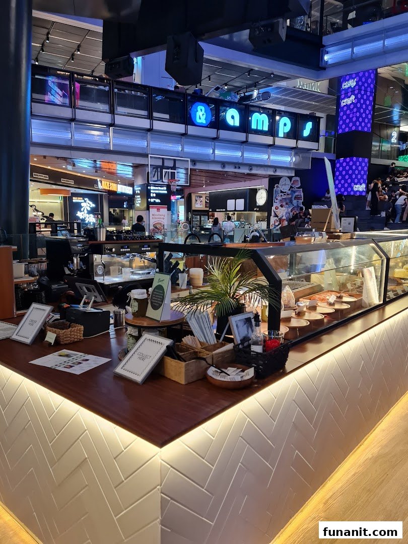 A vibrant coffee shop counter with a display case, plants, and various items, set within a bustling mall.