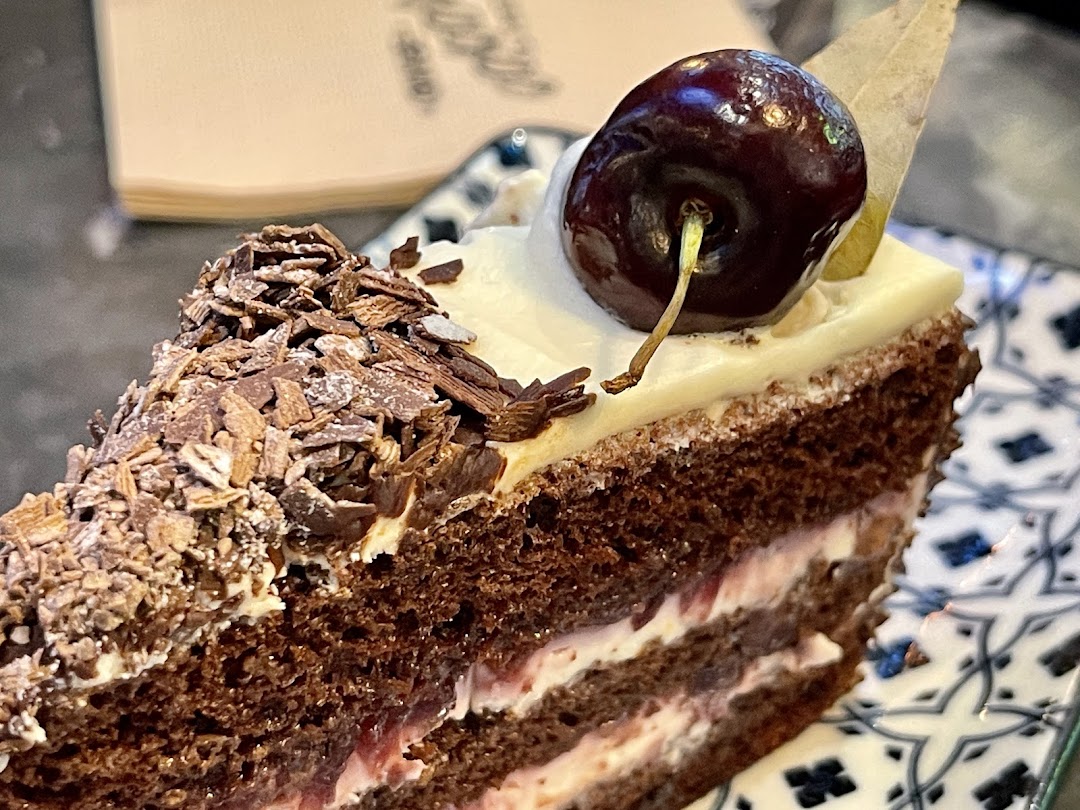 A close-up of a rich chocolate cake slice, garnished with a cherry and chocolate shavings.