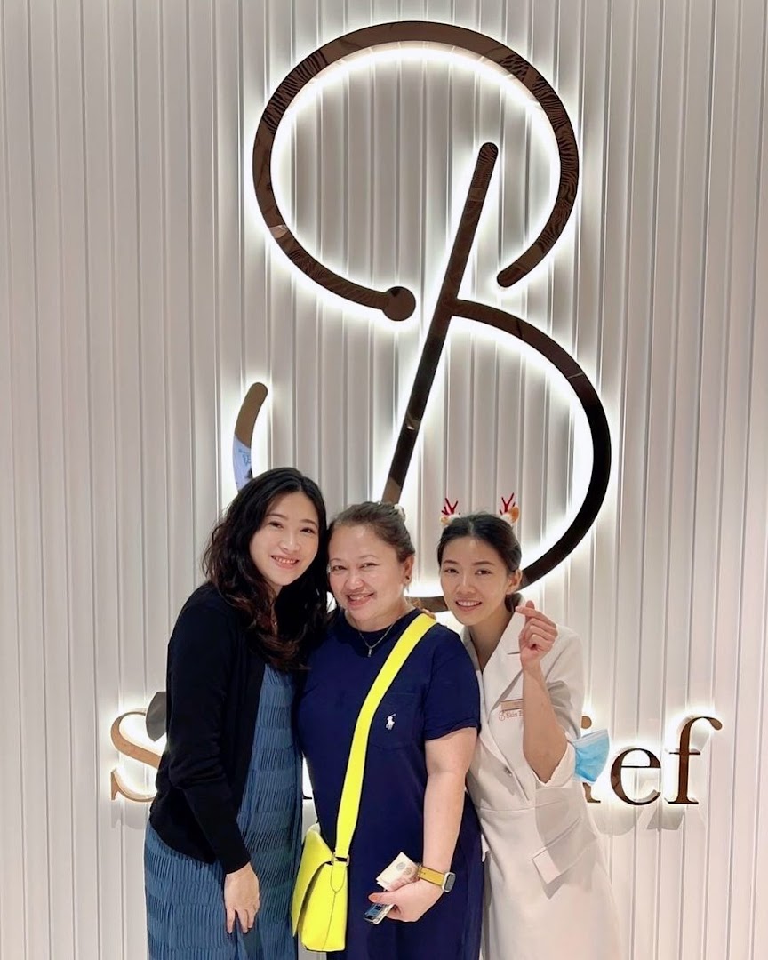 Three women smiling in front of the illuminated Skin Belief logo inside the shop.