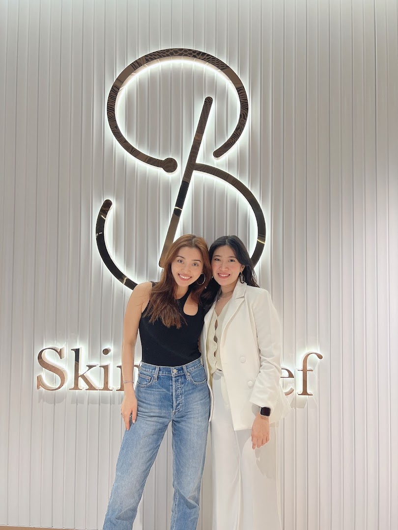 Two smiling women stand in front of the illuminated Skin Belief logo and brand name.