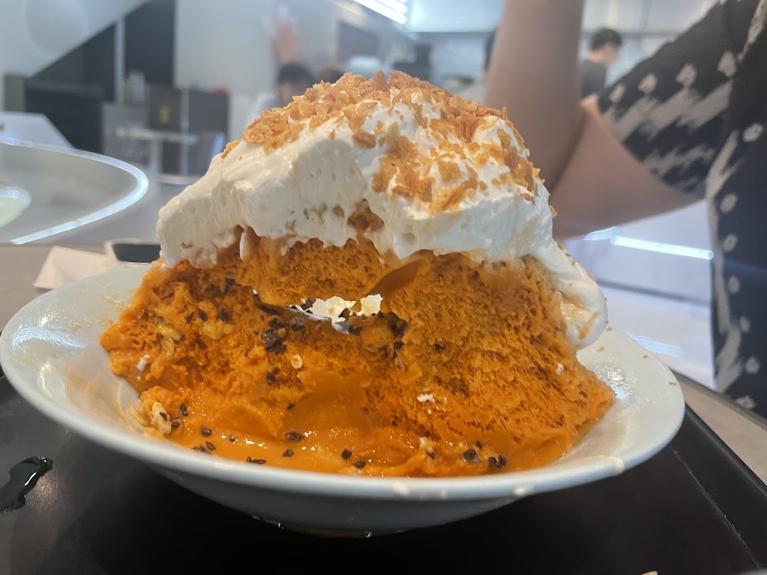 A large, vibrant orange shaved ice dessert topped with whipped cream and crunchy flakes.