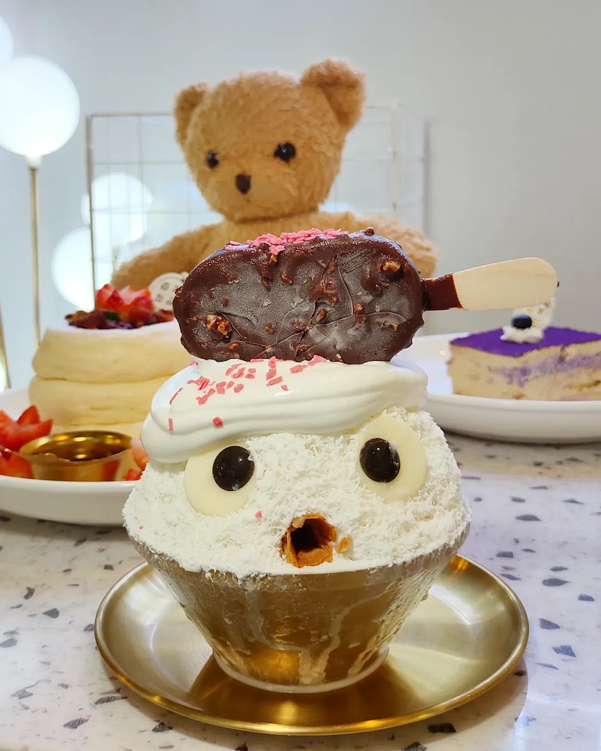 A whimsical dessert featuring a character-shaped shaved ice topped with an ice cream bar, alongside other treats and a teddy bear.