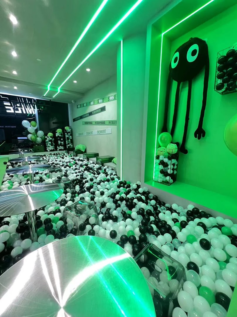 A vibrant, green-lit dessert shop interior featuring a large ball pit, modern tables, and playful decor.