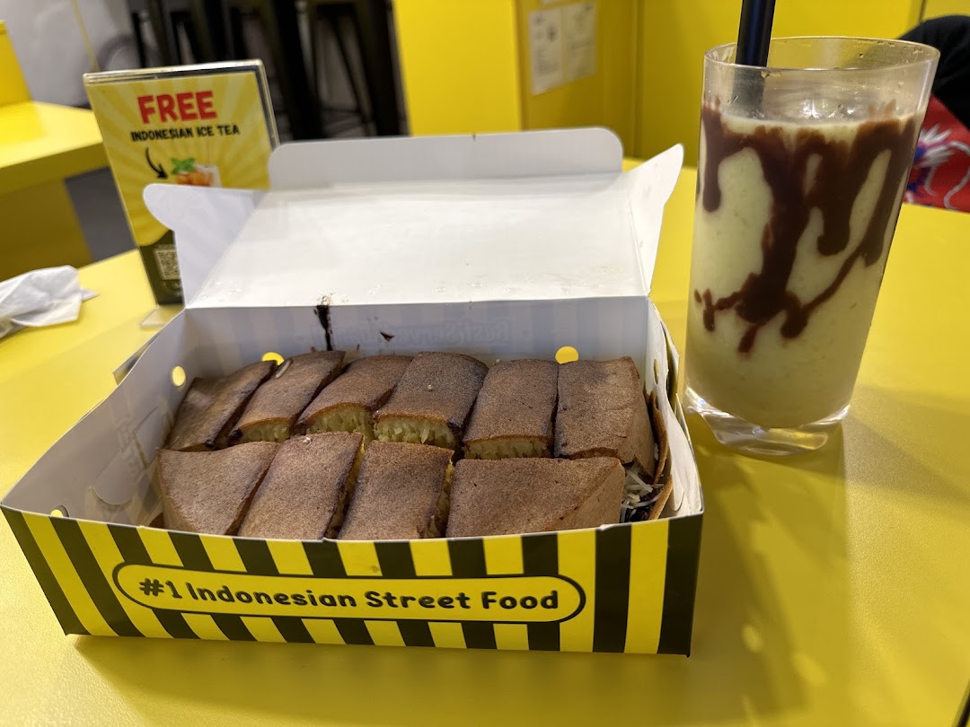 Delicious martabak slices in a box with a refreshing drink on a vibrant yellow table.