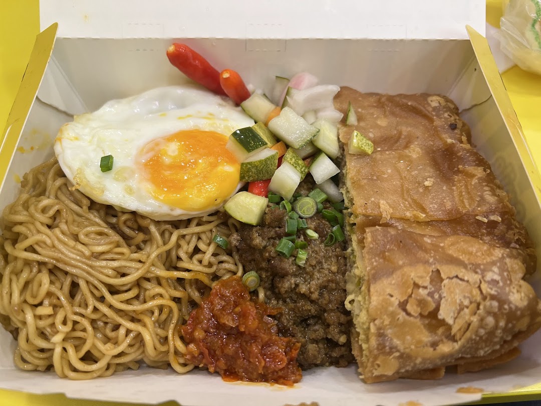 A takeaway box featuring noodles, a fried egg, minced meat, pickles, chili, and a piece of martabak.