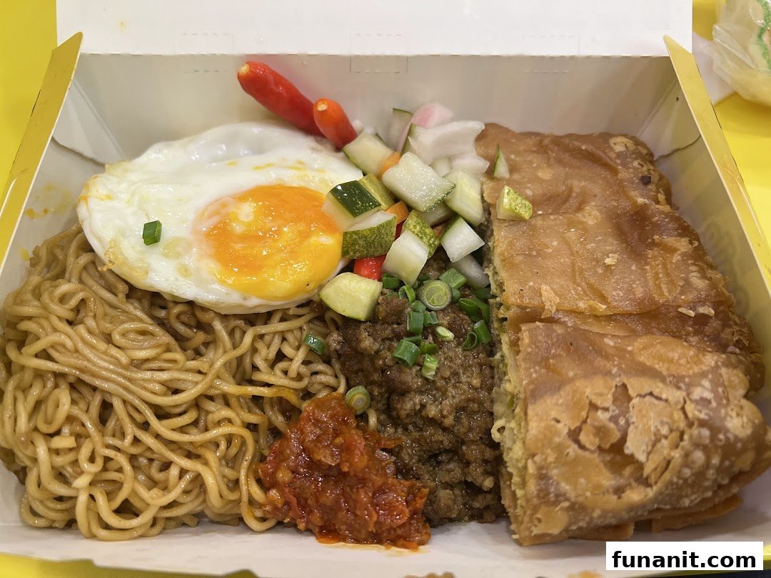 A takeaway box featuring noodles, a fried egg, minced meat, pickles, chili, and a piece of martabak.