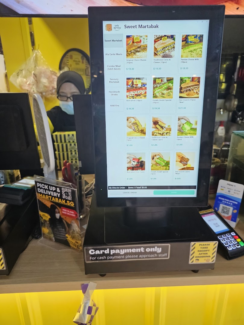 A digital menu display at a counter, showing various martabak options and prices, with a staff member in the background.