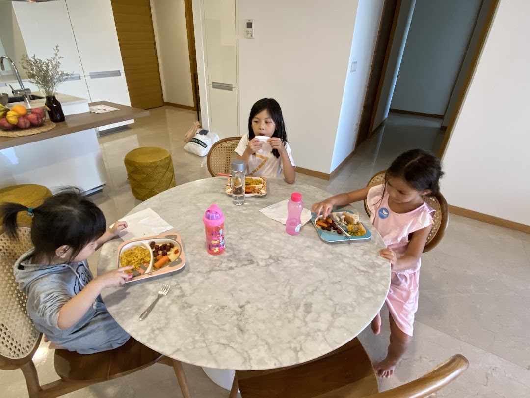 Three children are seated around a round table, eating meals in a bright home kitchen setting.